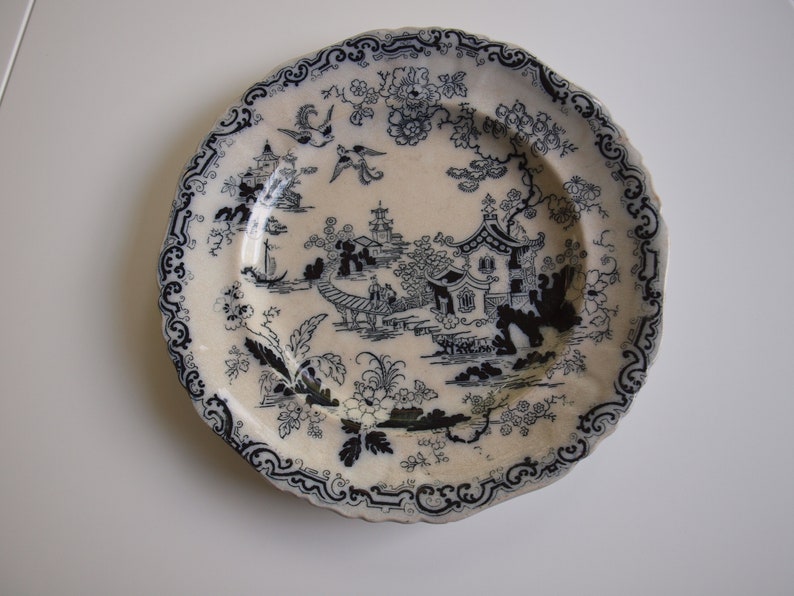 K&ouml;nnte beinhalten: Ein cremefarbener Teller mit einem blau-wei&szlig;en, asiatisch inspirierten Design. Der Teller zeigt eine detaillierte Landschaftsszene mit Pagoden, Br&uuml;cken und V&ouml;geln, umrahmt von einem dekorativen Rand. Ein dekorativer Teller im Vintage-Stil.
