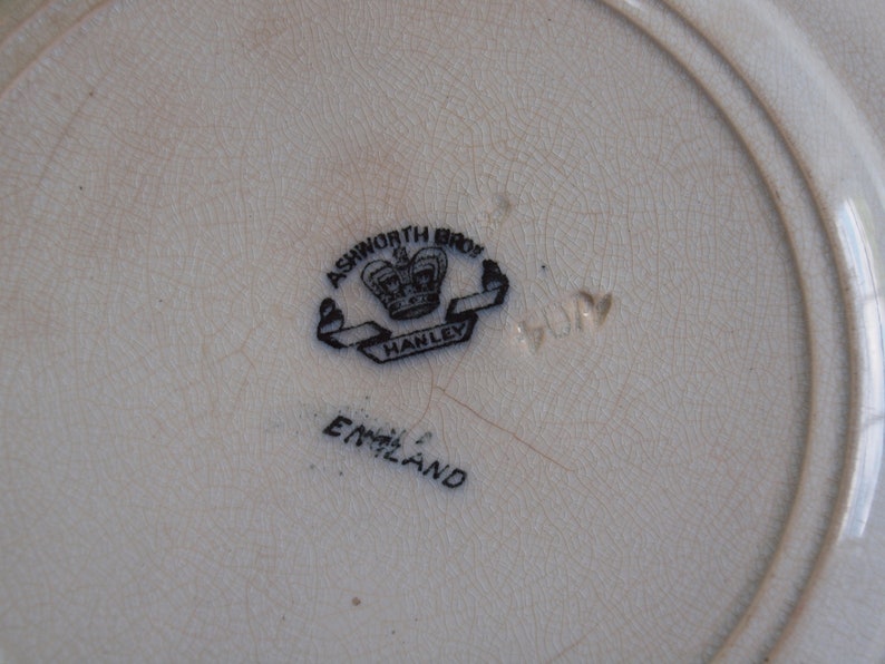 K&ouml;nnte beinhalten: Nahaufnahme eines cremefarbenen Vintage-Tellers mit rissiger Oberfl&auml;che. Der Teller zeigt ein schwarz-wei&szlig;es Emblem mit einer Krone und den Worten "ASHWORTH BROS HANLEY ENGLAND". Die Nummer "404" ist ebenfalls sichtbar.
