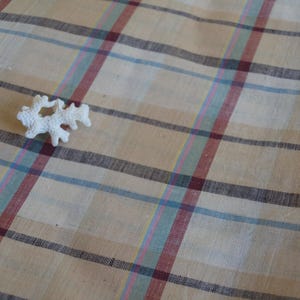 May include: A beige fabric featuring a plaid pattern with intersecting lines in shades of dark red, light blue, and green. A small, white, textured, branch-like coral piece is positioned on the surface of the woven textile material.