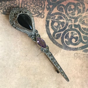 K&ouml;nnte beinhalten: Eine silberfarbene Vintage-Brosche mit Markasit-Akzenten. Die Brosche hat einen tropfenf&ouml;rmigen schwarzen Stein, einen ovalen Amethysten in der Mitte und ein langes, spitzes Design. Ein klassisches Schmuckst&uuml;ck.
