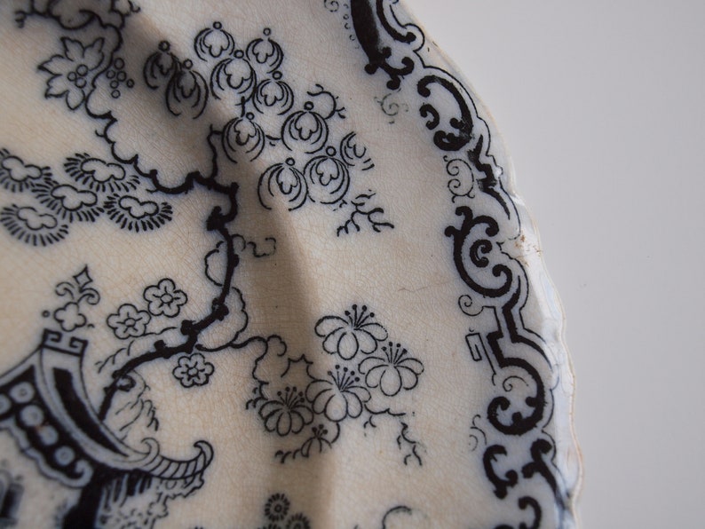 K&ouml;nnte beinhalten: Nahaufnahme eines wei&szlig;en Keramiktellers mit einem schwarzen floralen und architektonischen Design. Der Teller zeigt ein detailliertes Muster aus Bl&uuml;ten, Zweigen und einer Pagodenstruktur mit dekorativem Rand.