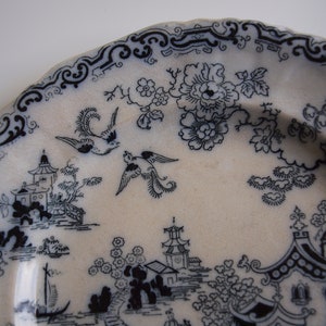 K&ouml;nnte beinhalten: Ein wei&szlig;er Keramikteller mit einem schwarz-wei&szlig;en, asiatisch inspirierten Design. Der Teller zeigt eine detaillierte Landschaft mit Pagoden, V&ouml;geln und floralen Mustern. Der Rand hat eine dekorative Bord&uuml;re.