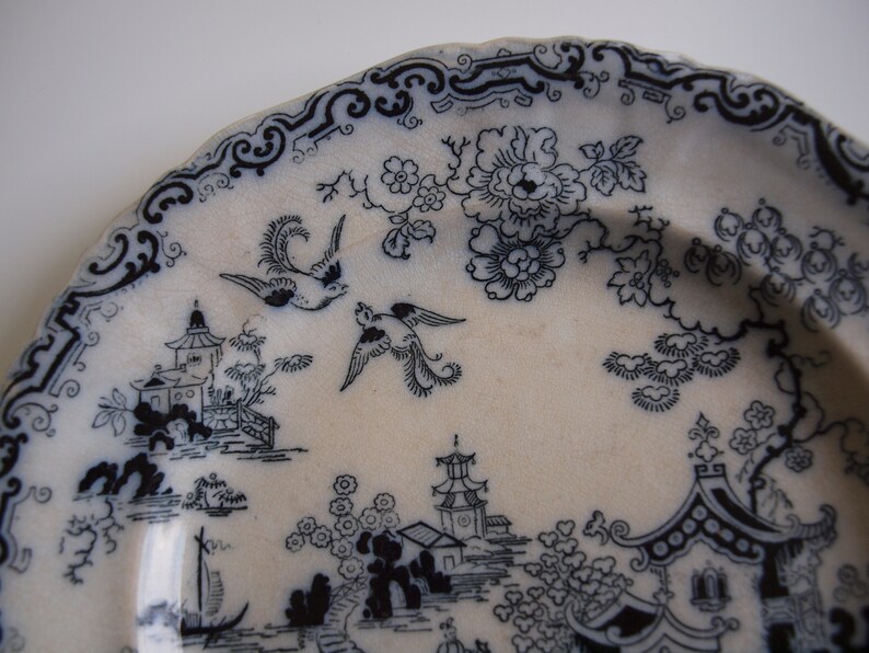 K&ouml;nnte beinhalten: Ein wei&szlig;er Keramikteller mit einem schwarz-wei&szlig;en, asiatisch inspirierten Design. Der Teller zeigt eine detaillierte Landschaft mit Pagoden, V&ouml;geln und floralen Mustern. Der Rand hat eine dekorative Bord&uuml;re.
