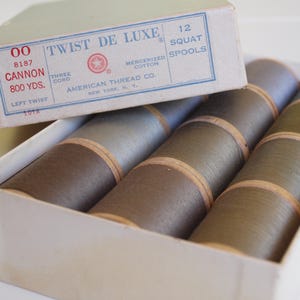 May include: A box of 12 squat spools of Cannon Twist De Luxe mercerized cotton thread. The box lid features the text "TWIST DE LUXE," "12 SQUAT SPOOLS," "CANNON," "800 YDS," and "AMERICAN THREAD CO." The spools are arranged in three rows of four.