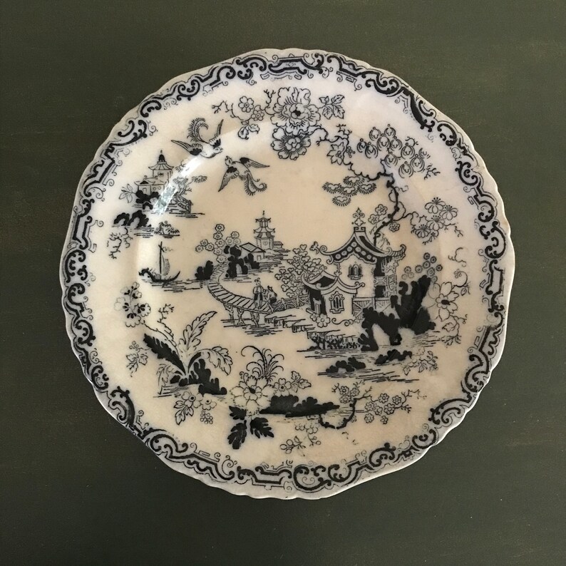 A white ceramic plate with a blue and white design of a pagoda and other Asian-inspired imagery. The plate has a scalloped edge and a blue floral pattern around the rim. It is from Staffordshire, ca 1890.