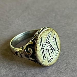 May include: A silver signet ring with an oval face featuring an engraved monogram of the letters "K" and "U". The ring has a decorative band with a floral design.