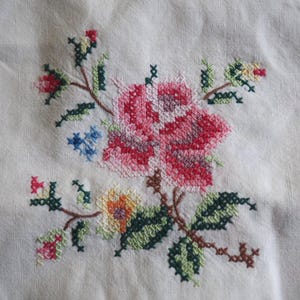 May include: Close-up of a white fabric with a cross-stitched floral design. The embroidery features a large pink rose, green leaves, and smaller flowers in blue, yellow, and red. The stitches are detailed and colorful.