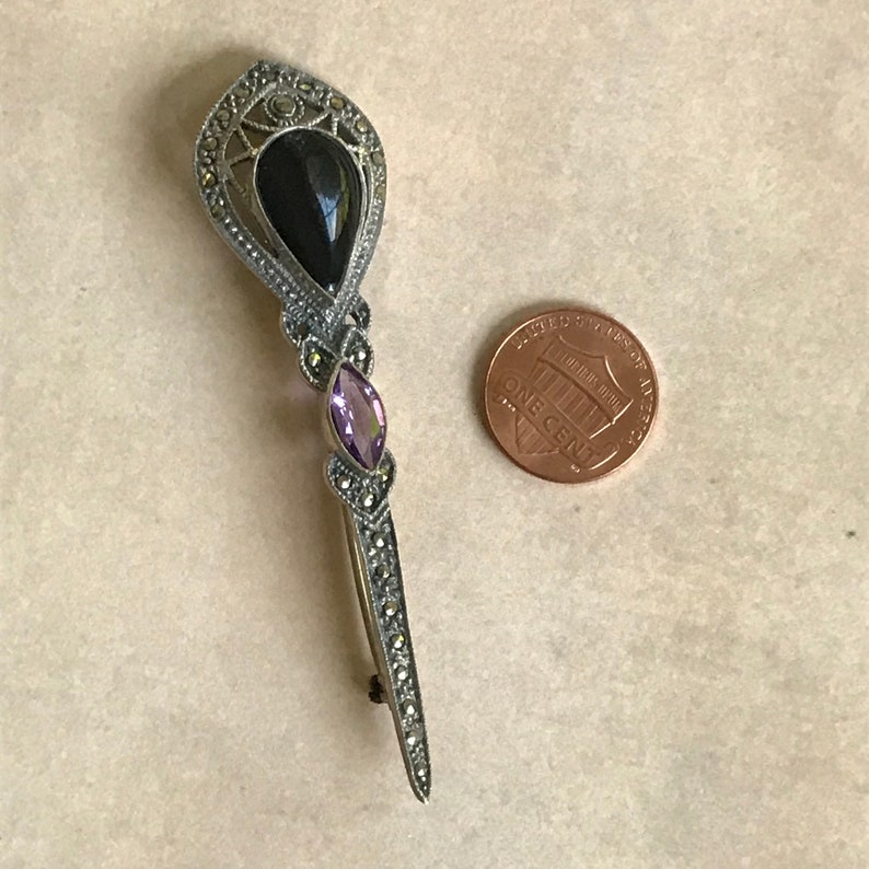 K&ouml;nnte beinhalten: Vintage Brosche in Silberoptik mit einem tropfenf&ouml;rmigen schwarzen Stein und einem ovalen lila Stein. Die Brosche ist mit kleinen, runden Markasitsteinen besetzt. Eine Ein-Cent-M&uuml;nze liegt daneben zum Gr&ouml;&szlig;envergleich.