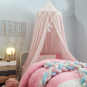 Puede incluir: Un dosel de cama de tela rosa pálido con parte superior puntiaguda y borde de pompones, sobre una cama con ropa de cama rosa y un cojín trenzado grueso azul y blanco. Un atrapasueños cuelga dentro, iluminado por luces de cadena.