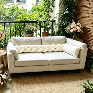 Può includere: Un divano a due posti beige chiaro con un lungo cuscino intrecciato color crema posizionato orizzontalmente sui cuscini della seduta, accompagnato da un cuscino rettangolare sul lato destro, il tutto davanti a un balcone con piante verdi.