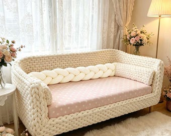 以下が含まれることがあります： フレームとアームにチャンキーニットの質感を施したクリーム色のソファ。座面は白の水玉模様が入った淡いピンク色の生地で覆われています。背もたれには、クリーム色の長い編み込み状のボルスタークッションが置かれています。