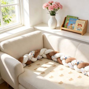 May include: A long, braided crib bumper featuring alternating segments of solid white, solid brown, and white fabric with a small bear print. The bumper rests on a cream-colored mattress pad decorated with a repeating pattern of small brown bear faces.