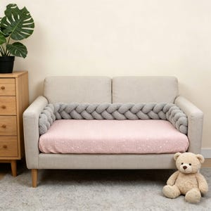 May include: A light beige sofa featuring a soft pink mattress cover with a subtle white star pattern and a thick, grey braided fabric bumper guard that wraps around the inner perimeter of the seating area, providing a cushioned border.