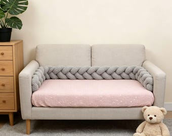 以下が含まれることがあります： 薄いベージュ色のソファ。ピンク色の柄入りマットレスカバーと、座面の内側を囲むように配置された長いグレーの編み込み式バンパークッションが特徴です。ソファはすっきりとしたフレームと明るい色の木製脚を備えています。