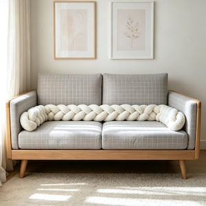Könnte beinhalten: Ein langes, cremefarbenes geflochtenes Nackenkissen mit einer weichen, strukturierten Oberfläche. Das Kissen liegt horizontal auf der Sitzfläche eines hellgrauen Sofas mit dezentem weißem Karomuster und einem hellen Holzrahmen.