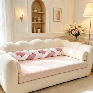 Puede incluir: Un cojín largo y trenzado con un patrón tejido de hebras de tela rosa suave y blanca. El cojín está colocado horizontalmente sobre el asiento de un sofá de color crema, cubierto con una colchoneta rosa claro con lunares blancos.