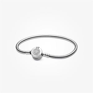 Op de afbeelding: Een zilverkleurige slangenkettingarmband met een ronde tonvormige sluiting. De sluiting is versierd met een cirkelvormige opstelling van heldere stenen en de gegraveerde tekst PANDORA. De armband heeft een flexibel, getextureerd schakelontwerp.