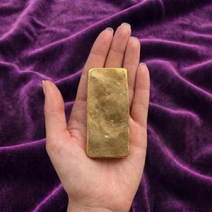 Puede incluir: Una barra rectangular de color dorado con una superficie texturizada e irregular. El objeto tiene esquinas redondeadas y un acabado metálico, mostrando una cualidad reflectante en su cara frontal. La barra es sólida y de tono dorado.