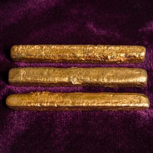 May include: Three rectangular gold-coloured bars with a rough, textured surface. Each bar has rounded edges and a metallic finish, arranged horizontally in a parallel stack. The bars exhibit a raw, uneven appearance with variations in the gold tone.