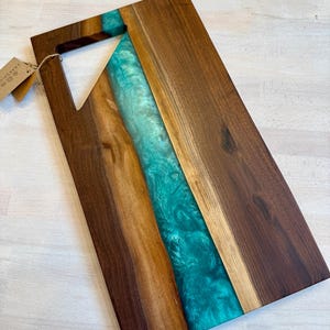 May include: A rectangular wooden cutting board with a turquoise resin river running down the center. The board has a handle cut out of one corner. The wood is a dark brown color, and the resin has a marbled effect.