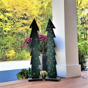 May include: Two wooden Christmas tree decorations with lights, standing on a wooden porch with a fall foliage background.