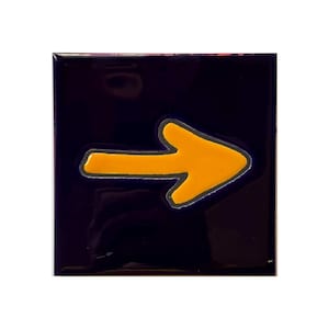 May include: A square tile with a glossy dark navy blue finish featuring a centered, bold yellow arrow pointing to the right. The arrow has a thin, light gray outline that separates the yellow shape from the dark blue background of the tile.