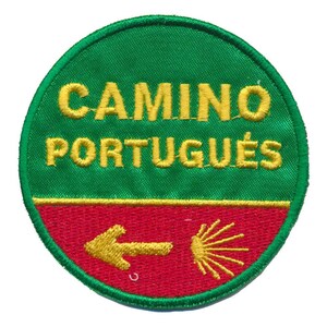 Op de afbeelding: Een cirkelvormige geborduurde patch, groen boven en rood beneden, met gele geborduurde tekst "CAMINO PORTUGUÉS". Een gele pijl en een gestileerde gele schelp zijn ook op de patch geborduurd.