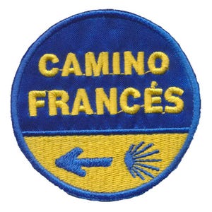 Op de afbeelding: Een ronde, geborduurde patch met de tekst "CAMINO FRANCÉS" in gele draad op een blauwe achtergrond. De onderste helft is geel met een blauwe pijl en een gestileerde sint-jakobsschelp. De patch is afgewerkt met blauwe stiksels.