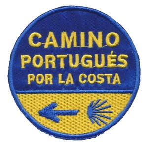 Op de afbeelding: Ronde patch met gele borduursels op een blauwe achtergrond. De tekst is "CAMINO PORTUGUÊS POR LA COSTA". Een gele sectie aan de onderkant toont een blauwe pijl en een gestileerde sint-jakobsschelp.