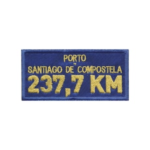 Może przedstawiać: Prostokątna haftowana naszywka z ciemnoniebieskim tłem i napisami wykonanymi złotą nicią. Tekst brzmi PORTO SANTIAGO DE COMPOSTELA 237,7 KM. Naszywka posiada obszyte brzegi i jest przeznaczona do umieszczenia na tkaninach lub odzieży.