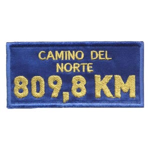 May include: A rectangular blue patch with gold embroidered text. The text reads "CAMINO DEL NORTE" above "809.8 KM". The patch has a blue border and is likely a souvenir or emblem.