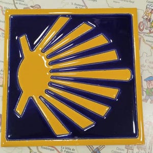 May include: A square ceramic tile featuring a glossy dark blue background with a raised, bright yellow scallop shell emblem. The shell design consists of a central semi-circle with radiating lines extending toward the right edge of the tile.