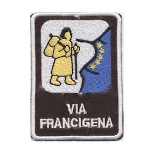 Può includere: Una toppa ricamata rettangolare con bordo marrone scuro e rifinitura bianca. Al centro presenta una figura gialla di un escursionista con zaino e bastone, una sezione blu con stelle gialle e il testo bianco VIA FRANCIGENA in basso.