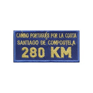 Op de afbeelding: Een rechthoekige blauwe geborduurde patch met gouden tekst die luidt CAMINO PORTUGUÉS POR LA COSTA SANTIAGO DE COMPOSTELA 280 KM. De patch heeft een gestikte rand en een gestructureerd stoffen oppervlak met duidelijke, dikgedrukte letters.