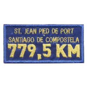 Op de afbeelding: Een rechthoekige blauwe geborduurde patch met gouden tekst. De tekst luidt "ST. JEAN PIED DE PORT SANTIAGO DE COMPOSTELA 779,5 KM". De patch heeft een blauwe rand.