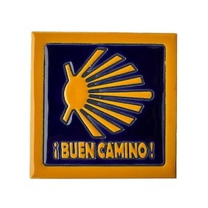 May include: A square ceramic tile featuring a glossy dark blue center with a yellow scallop shell emblem and the yellow text ¡BUEN CAMINO! printed below. The tile is framed by a solid yellow border, creating a vibrant and high-contrast design.