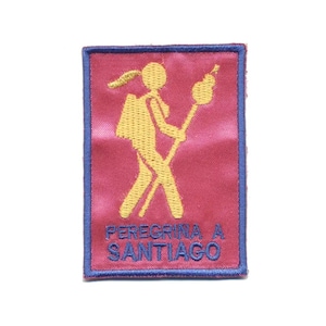 May include: A rectangular embroidered patch with a deep pink background and a blue border. It features a yellow stick figure carrying a backpack and a staff with a gourd. The blue text PEREGRINA A SANTIAGO is embroidered at the bottom.