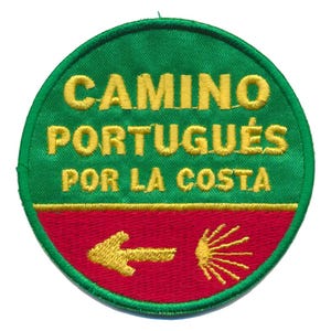 Op de afbeelding: Een ronde, geborduurde patch met de tekst "CAMINO PORTUGUÉS POR LA COSTA" in geel op een groene achtergrond. Het onderste gedeelte is rood, met een gele pijl en een gestileerd schelpsymbool.
