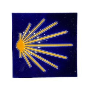 May include: A square ceramic tile with a glossy deep blue finish featuring a stylized yellow scallop shell emblem. The design consists of a central semi-circular shape with radiating lines extending outward, creating a sunburst pattern across the surface.