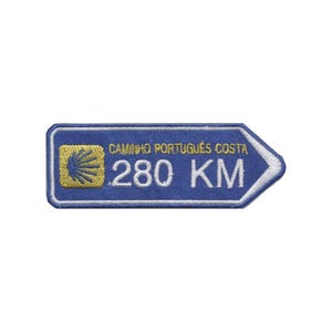 May include: A blue embroidered patch shaped like a directional arrow. It features a yellow scallop shell icon on the left, the text CAMINHO PORTUGUÊS COSTA in yellow, and the white text 280 KM, all outlined with a white border on a blue background.