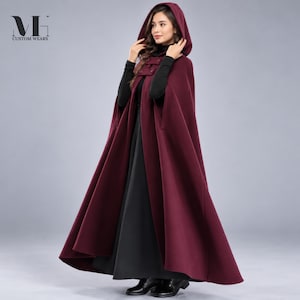 May include: A floor-length, deep burgundy hooded cloak featuring a structured collar with two visible buttons at the neck. The garment is worn over a black long-sleeved top and a black skirt. The logo MH CUSTOM WEARS is visible in the top left corner.