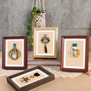 Puede incluir: Cuatro medallas enmarcadas en marcos de madera con paspartú beige. Una medalla es circular con un edificio, una es un candado en forma de corazón con borla, una es un cuadrado de filigrana dorada y una es una medalla en forma de estrella.