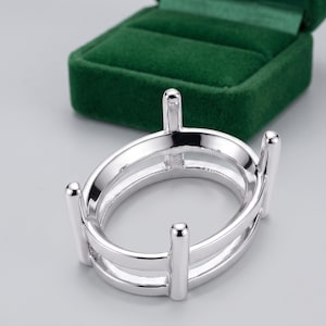 May include: A polished silver-tone oval ring setting featuring a double-band design and four vertical prongs extending upward to secure a gemstone. The metal surface is smooth and reflective, showcasing a clean, structural frame for jewelry.
