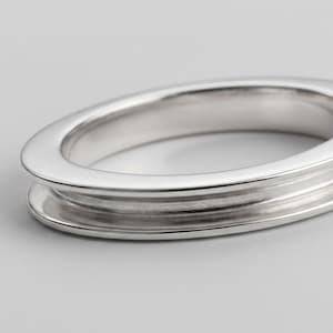 Könnte beinhalten: Ein polierter Silberring mit einem glatten, abgerundeten Außenband und einer ausgeprägten, vertieften Rille in der Mitte. Das Metall hat eine reflektierende Oberfläche, eine klare, durchgehende Kreisform und eine einheitliche Konstruktion.