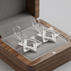 May include: A pair of silver earrings featuring a six-pointed star design with a hollow center and curved ear wires. The earrings are displayed inside a wooden box with a grey velvet interior lining, showcasing a polished metallic finish.