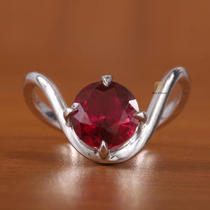 May include: A silver-toned ring featuring a round, faceted deep red gemstone held by four prongs. The band has a smooth, curved, wave-like design that rises to meet the central stone. A small logo with the text "Twist & Forever" is visible.
