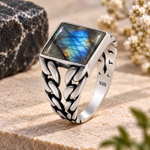 May include: A sterling silver ring featuring a square-cut labradorite gemstone with iridescent blue and gold flashes. The band is designed with a detailed chain-link pattern on both sides and is clearly stamped with the 925 hallmark inside.