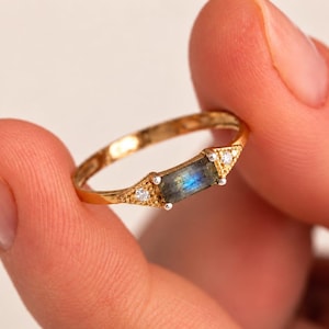 May include: A gold ring featuring a horizontal baguette-cut labradorite gemstone with a blue flash, flanked by two small round white stones set in triangular textured gold settings on either side of the central stone on a thin gold band.