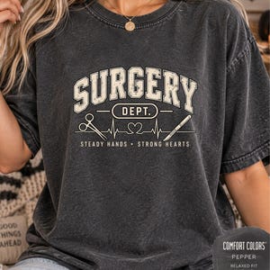 Könnte beinhalten: Ein anthrazitfarbenes Kurzarm-T-Shirt mit dem Text SURGERY DEPT. in einer cremefarbenen College-Schriftart. Unter dem Text befindet sich eine Grafik mit einer chirurgischen Schere, einer herzförmigen EKG-Linie und einem Skalpell mit STEADY HANDS STRONG HEARTS.