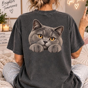 Puede incluir: Una camiseta de manga corta gris carbón con un acabado lavado vintage. La parte posterior de la camiseta presenta un gráfico grande y detallado de un gato gris con ojos de color ámbar brillante, mostrado desde el pecho con las patas delanteras.